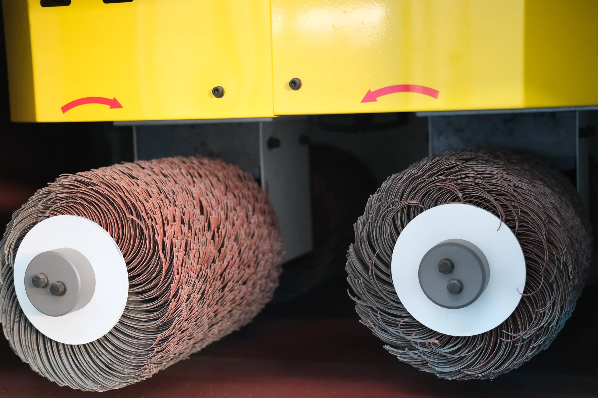 Two abrasive brush rollers beneath a yellow guard with rotation-direction arrows.